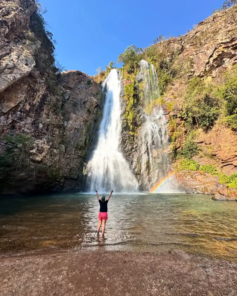 Visitante de braços abertos diante de cachoeira com arco-íris